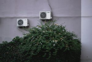 photo of air conditioning units attached to an exterior wall with a green bush underneath